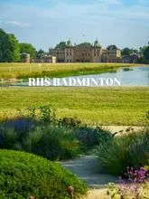 A bright, clear landscape photograph of Badminton House, serving as the backdrop for the RHS Flower Show. The composition features a garden path in the foreground leading the eye toward the historic estate and a lake.
