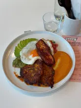 A top-down, flat-lay photograph of a brunch dish featuring sweetcorn fritters, a fried egg, avocado, and halloumi, served on a white plate at a cafe.