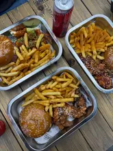 A top-down, flat-lay photograph of three metal trays containing burgers, fries, and chicken wings, served on a wooden table. The image captures a casual dining experience, likely at a burger or street food venue.