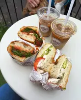 A casual, overhead shot of two sandwiches and two iced coffees on a white table, likely taken at a cafe.