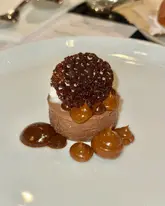 A close-up, top-down shot of a single, small-portion dessert from a tasting menu, featuring a chocolate mousse base topped with a dark tuile, served with dollops of caramel or sauce on a white plate.