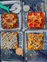 A top-down, symmetrical flat-lay photograph of four Detroit-style square pizzas served on metal trays, each accompanied by a pizza cutter. The pizzas feature various toppings including pepperoni, jalapeños, mushrooms, and chicken, presented in a clean, casual dining setting.