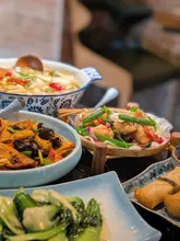 A top-down/angled shot of a spread of Sichuan dishes at a restaurant, featuring a large bowl of pickled cabbage and chilli fish, tofu, salt and pepper chicken, pak choi, and spring rolls. The lighting is standard indoor restaurant lighting, and the table is quite crowded with dishes.