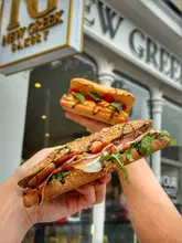 A bright, high-quality promotional image featuring two fresh sandwiches held up in front of a Greek bakery storefront. The focus is sharp on the foreground sandwich, effectively showcasing the ingredients and texture, while the background provides clear context for the venue.