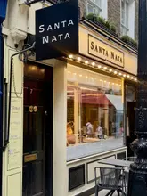 A vertical shot of the Santa Nata shop front in Covent Garden, capturing the warm, inviting exterior of the Portuguese bakery. The image focuses on the signage and the window display, conveying a cozy, authentic atmosphere.
