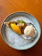 A high-quality, top-down shot of a single oyster dish served on a bed of rock salt in a ceramic bowl, set against a wooden table. The dish features precise, artistic plating with a vibrant yellow element, a piece of seafood, and a creamy foam, showcasing the fine dining nature of the venue.