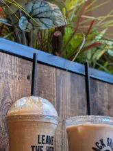 A close-up, eye-level shot of two plastic coffee cups from Black Sheep Coffee, set against a wooden wall with some greenery visible in the background.
