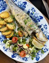 A high-quality, overhead shot of a whole grilled seabass served with sliced potatoes and a fresh salad on a patterned plate, likely taken on an outdoor terrace.