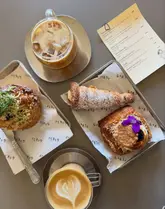 A high-quality, overhead flat-lay photograph of a selection of artisanal pastries and coffee from a bakery. The image features a tray with a 'Tayto' pastry, a 'Fifteen' Danish, and a Pump Street cream horn, alongside an iced coffee and a cappuccino with latte art. A printed menu is visible on the table, providing context for the items.