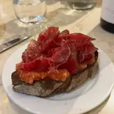 A close-up, eye-level shot of a single slice of toasted bread topped with tomato and thin shavings of cured cecina meat, served on a white plate.