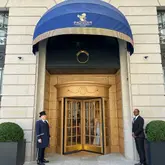 A high-quality, symmetrical exterior shot of The Ritz London entrance, capturing the iconic blue awning, gold detailing, and the hotel's doormen. The image effectively conveys the prestige and luxury associated with the venue.