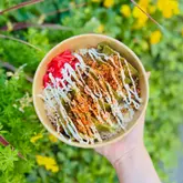 A high-quality, overhead shot of a Japanese gyoza rice bowl held in a hand against a blurred natural green background. The dish is well-presented with a generous drizzle of mayonnaise, crispy toppings, and pickled ginger, making it look fresh and appealing.