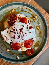 A high-quality, overhead shot of a brunch dish featuring a poached egg, roasted cherry tomatoes, and pine nuts on toast, drizzled with oil and chili flakes.