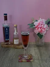 A simple, home-style photograph showcasing a cocktail made with blackberry gin liqueur and pink prosecco. The image features the ingredients and the finished drink on a wooden surface against a plain pink wall.