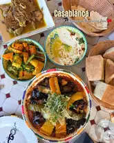 A vibrant, well-composed overhead flat-lay shot of a Moroccan feast, featuring lamb tangia, moussaka tagine, hummus, and fresh bread, set against a patterned table.