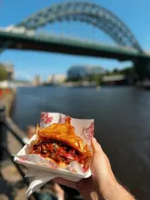A high-quality, appetizing close-up shot of a barbacoa beef torta held in hand, set against the iconic backdrop of the Tyne Bridge in Newcastle. The image effectively uses a shallow depth of field to isolate the food while providing clear location context.