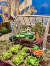 A high-quality, well-composed photograph of a Wallace & Gromit model set from the 'Inside Aardman' exhibition at the Young V&A museum. The image captures the intricate detail of the miniature greenhouse and garden scene, showcasing the craftsmanship of the Aardman animation team.