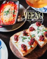 A high-quality, well-lit promotional image featuring Italian small plates and wine, likely for a wine bar or restaurant. The composition focuses on a piece of focaccia topped with cheese and pesto, with a baked dish and a glass of wine in the background.
