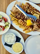A top-down, natural-light shot of a Greek mixed grill platter featuring skewers, fries, and pitta, served alongside a side salad and two dipping sauces on a wooden table.