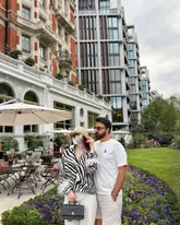 A lifestyle photograph featuring a couple posing in front of an upscale hotel, likely captured for social media content. The image focuses on the aesthetic of the venue and the couple's attire.
