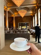 A first-person perspective shot of a latte with latte art, held in hand, with the opulent, warm-lit interior of the RH (Restoration Hardware) London venue in the background.