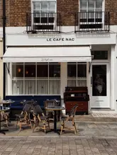 A straight-on architectural shot of the exterior of Le Café Nac in Belgravia, London. The image captures the cafe's storefront, featuring outdoor seating with bistro-style chairs and tables, a white awning, and a brick facade. The lighting is soft and overcast, typical of a cloudy day.