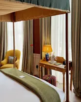 A beautifully composed, high-quality photograph of a boutique hotel room at The Zetter Bloomsbury. The image captures a serene, inviting atmosphere with a four-poster bed in the foreground and a desk area featuring a champagne bucket, showcasing the elegant and comfortable interior design.