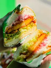 A high-contrast, close-up macro shot of a vegan burger held in hand, showcasing the layers of the sandwich including crispy patty, lettuce, and pickled onions.