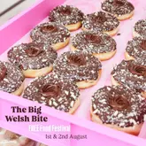 A promotional graphic for 'The Big Welsh Bite' food festival, featuring a repeating pattern of chocolate-topped donuts on a pink tray as the background for the event title and dates.