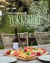 A promotional image for a luxury barn getaway in Yorkshire, featuring a grazing board and wine set on an outdoor table in a garden setting.