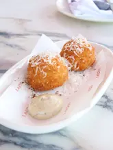 A high-quality, well-lit close-up shot of two parmesan truffle arancini served on branded paper on a marble table. The image captures the texture of the fried exterior and the grated cheese topping, making the dish look appealing and professional.
