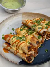 A high-quality, appetizing close-up shot of homemade taquitos topped with a creamy sauce and fresh cilantro, accompanied by a side of avocado cream. The image captures the texture and freshness of the dish effectively.