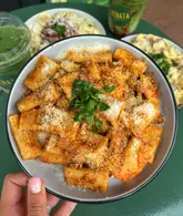 A top-down, handheld shot of a bowl of rigatoni pasta with a red sauce, topped with parmesan and parsley. The background features other dishes and green drinks, suggesting a casual dining experience at a food hall.
