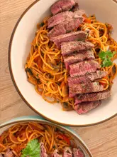 A high-quality, well-lit overhead shot of a bowl of spaghetti topped with sliced steak. The lighting is bright and natural, highlighting the texture of the meat and the sauce on the pasta. The composition is clean and focused, making the dish look appealing and well-prepared.