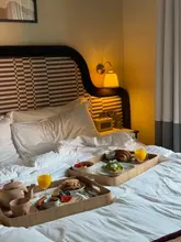 A high-quality, inviting lifestyle shot of a breakfast spread served on trays in a hotel bed. The warm, soft lighting creates a cozy and aspirational atmosphere, perfectly capturing the 'breakfast in bed' experience. The composition is well-balanced, with the food clearly presented and the setting feeling authentic and luxurious.