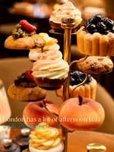 A high-quality, professional-grade photograph showcasing an elaborate afternoon tea stand featuring intricate, fruit-shaped French pastries. The image uses a shallow depth of field to isolate the subject against a warm, blurred background, emphasizing the craftsmanship of the food.