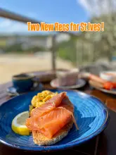 A bright, high-quality shot of a smoked salmon and scrambled egg breakfast dish served on toast, captured on a sunny terrace. The image uses a shallow depth of field to focus on the food while hinting at a scenic, outdoor seaside setting.