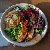 A top-down, eye-level shot of a takeaway Buddha bowl from Pret A Manger, featuring chicken, edamame, purple rice, and various vegetables, served in a disposable paper bowl on a wooden table.
