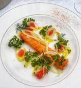 A high-quality, overhead shot of a beautifully plated salmon dish, likely a starter, served at The Cross at Kenilworth. The presentation is artistic, featuring fresh herbs, citrus segments, and shaved fennel, set against a clean, branded tablecloth.