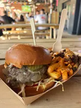 A candid, eye-level shot of a smash burger and loaded fries served in a cardboard tray, captured at a busy food hall venue.