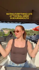 A creator reviews various dishes at The Real Greek in Bournemouth, rating each item from a mojito to a custard pie while seated at an outdoor table.