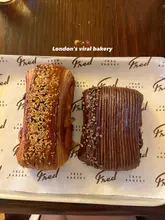 A high-quality, top-down photograph of two distinct pastries served on branded parchment paper. The image clearly showcases the texture and toppings of the baked goods, providing an appetizing view of the product.