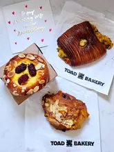 A flat-lay photograph of three assorted pastries from Toad Bakery, arranged on crumpled paper bags alongside an anniversary card on a white marble surface.