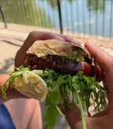 A close-up, handheld shot of a porchetta sandwich being held outdoors near a body of water. The image captures a casual, on-the-go dining moment, focusing on the texture of the bread and the filling.