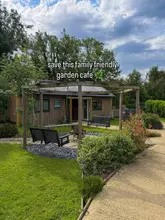A wide-angle, eye-level shot of an outdoor garden cafe setting, featuring wooden structures, hanging bench swings, and a central fire pit area, set in a lush, green environment under a partly cloudy sky.