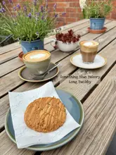 A high-angle, natural light shot of a salted caramel and pecan cookie served on branded paper, accompanied by two coffees on a rustic wooden table, set against a backdrop of potted plants and a brick wall.