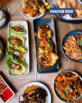 A high-quality, overhead flat-lay photograph showcasing a variety of Thai dishes, including prawn crackers, lettuce wraps, and stir-fried items, presented on a wooden table.