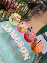 A vibrant, high-angle shot featuring four distinct spritz cocktails arranged in a diagonal line across a rustic Jameson-branded wooden barrel. The background is softly blurred, suggesting an outdoor pub or bar setting, which keeps the focus entirely on the colourful drinks.