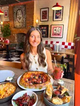A bright, inviting promotional shot featuring a diner posing with a table full of Italian-inspired dishes, including pizza, pasta, and cocktails, in a vibrant, decorated restaurant setting.