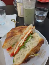 A close-up, top-down shot of a halloumi and roasted pepper focaccia sandwich served on branded paper, accompanied by an iced matcha latte. The lighting is natural and bright, highlighting the texture of the bread and the freshness of the ingredients.
